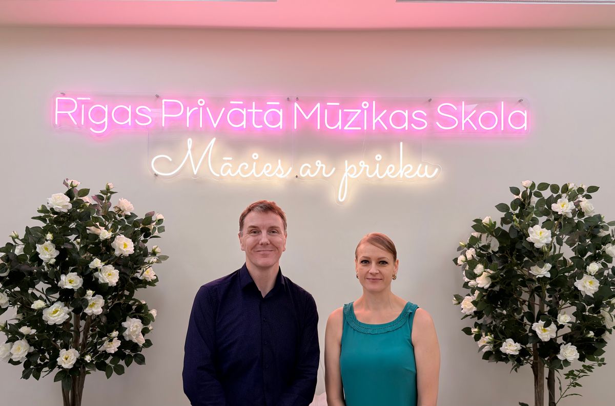 Profesors Pols Boids un Linda Boida, Rīgas Privātās Mūzikas Skolas dibinātāji, stāv zem skolas neona izkārtnes "Rīgas Privātā Mūzikas Skola — Mācies ar prieku"